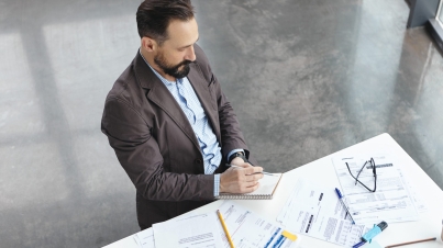 Top view of handsome skilled professional employer dressed formally, sits at work place, surrounded with many documents, writes with pencil in papers. Male marketing specialist works indoors